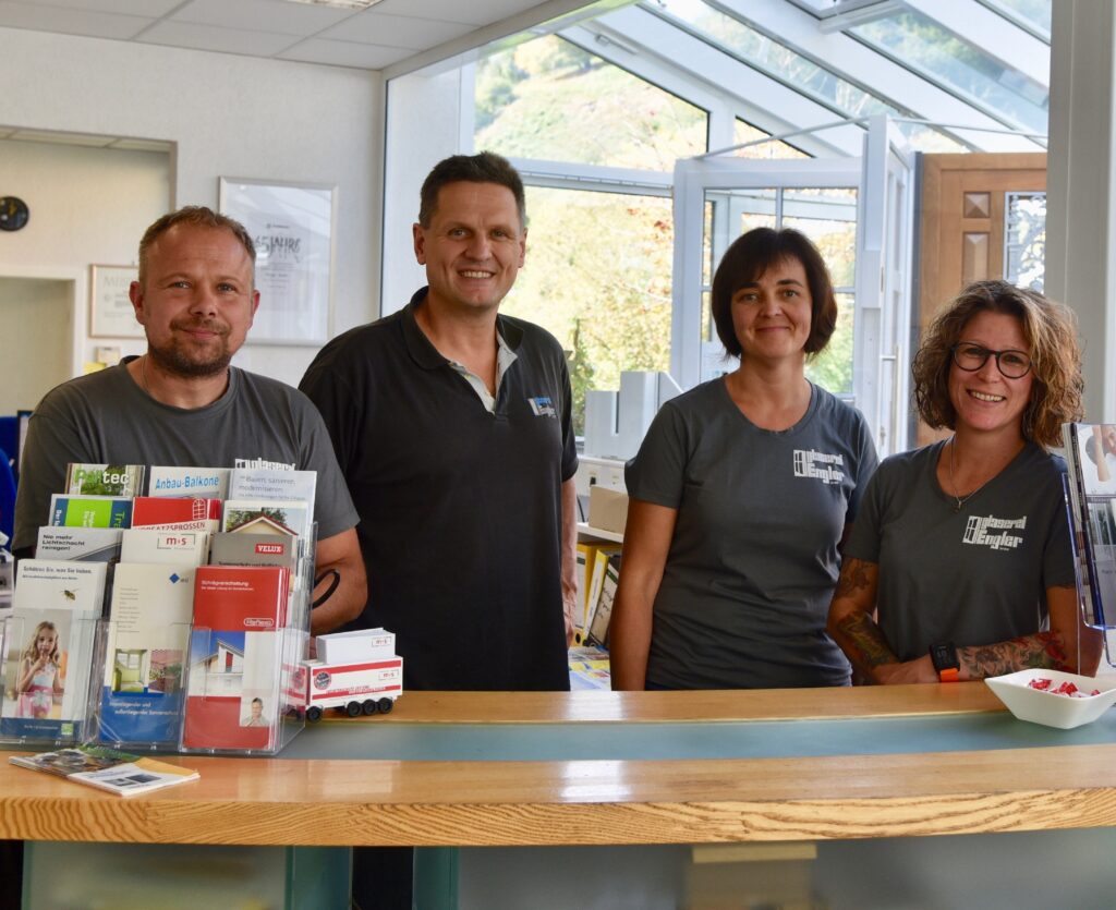 Das Glaserei-Büroteam, Zwei Männer und zwei Frauen stehen hinter einem Holztresen mit Ausstellern