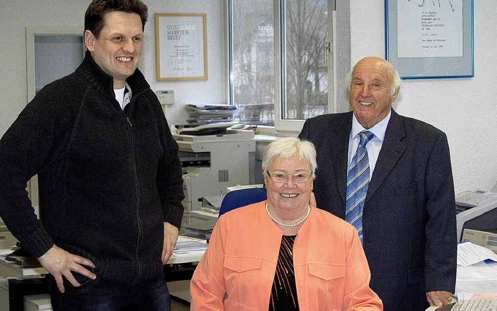 Zwei Männer im Anzug und eine Frau in lachsfarbenem Jacket lachen in eine Kamera